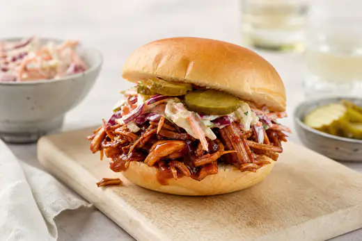 Weiches Burgerbrötchen mit Pulled Pork in BBQ-Sauce, belegt mit Krautsalat und Gurkenscheiben, auf einem Holzbrett