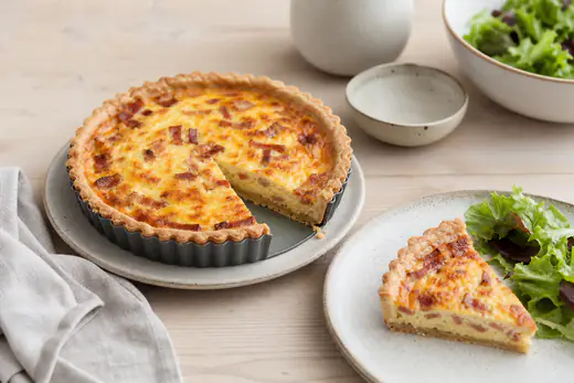 Kleine runde Quiche Lorraine mit Speck in der Tarteform, ein Stück auf einem Teller serviert, dazu grüner Salat