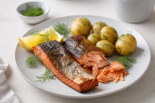 Warm geräucherte Lachsstücke mit Haut auf einem Teller mit Zitronenspalten, frischem Dill und gekochten neuen Kartoffeln