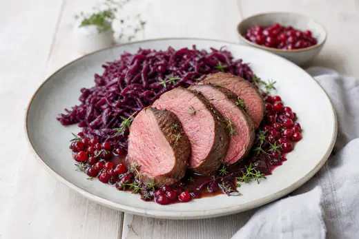 Rosa gebratener Rehrücken in dicke Scheiben geschnitten auf einem Teller, dazu Rotkohl, Preiselbeeren und Thymian