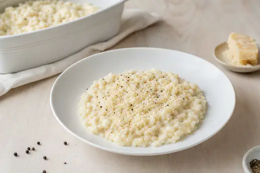 Cremiges weißes Risotto mit geriebenem Parmesan und schwarzem Pfeffer auf einem tiefen Teller, dahinter eine Auflaufform mit Risotto