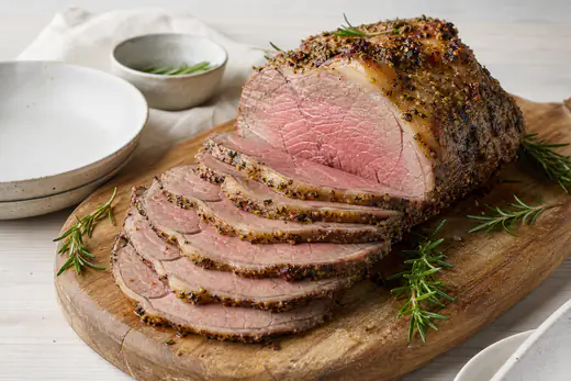 Roastbeef mit Fettkappe und Kräuter-Senf-Kruste in dünne rosa Scheiben geschnitten, auf einem Holzbrett mit Rosmarin