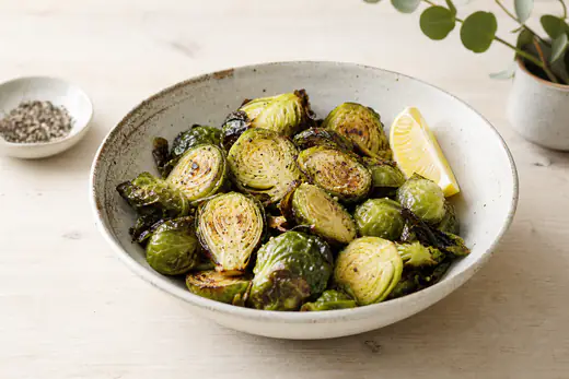 Halbierter gerösteter Rosenkohl mit dunklen knusprigen Blättern in einer Schüssel, mit Zitronenspalte und schwarzem Pfeffer
