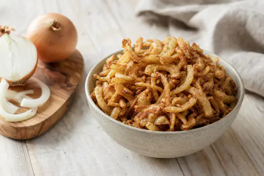 Kleine Schale mit goldbraunen knusprigen Röstzwiebeln, daneben rohe Zwiebeln auf einem Holzbrett