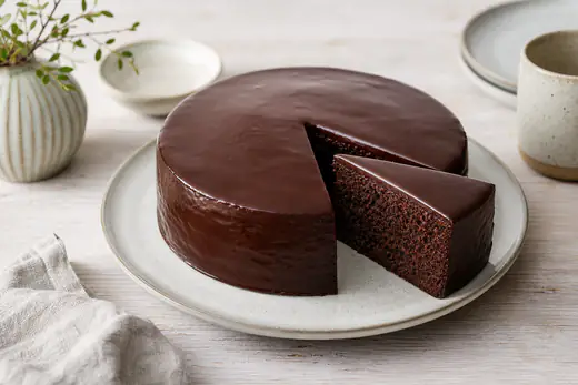 Kleiner runder Schokoladenkuchen mit glänzender dunkler Glasur auf einem Kuchenteller, ein Stück angeschnitten