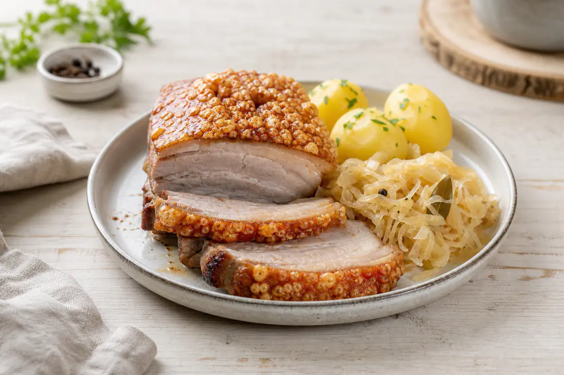 Aufgeschnittener Schweinebauch mit goldbrauner aufgeplatzter knuspriger Schwarte auf einem Holzbrett, dazu Sauerkraut und Kartoffeln