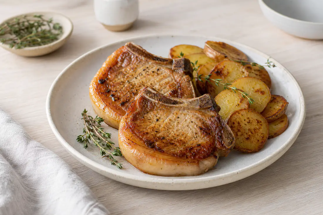 Zwei goldbraun gebratene Schweinekoteletts mit Knochen und knusprigem Fettrand, dazu Bratkartoffeln und Thymian