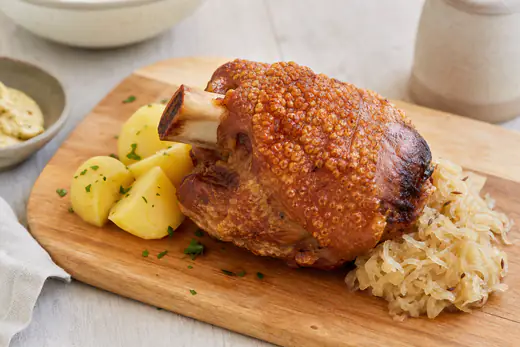 Eine goldbraune Schweinshaxe mit aufgeplatzter knuspriger Schwarte auf einem Holzbrett, daneben Sauerkraut und Salzkartoffeln