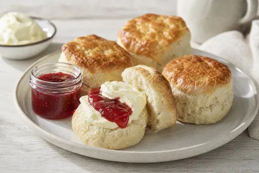 Goldbraune Scones, einer aufgeschnitten und mit Clotted Cream und Himbeermarmelade gefüllt, dazu weitere Scones und ein Marmeladenglas