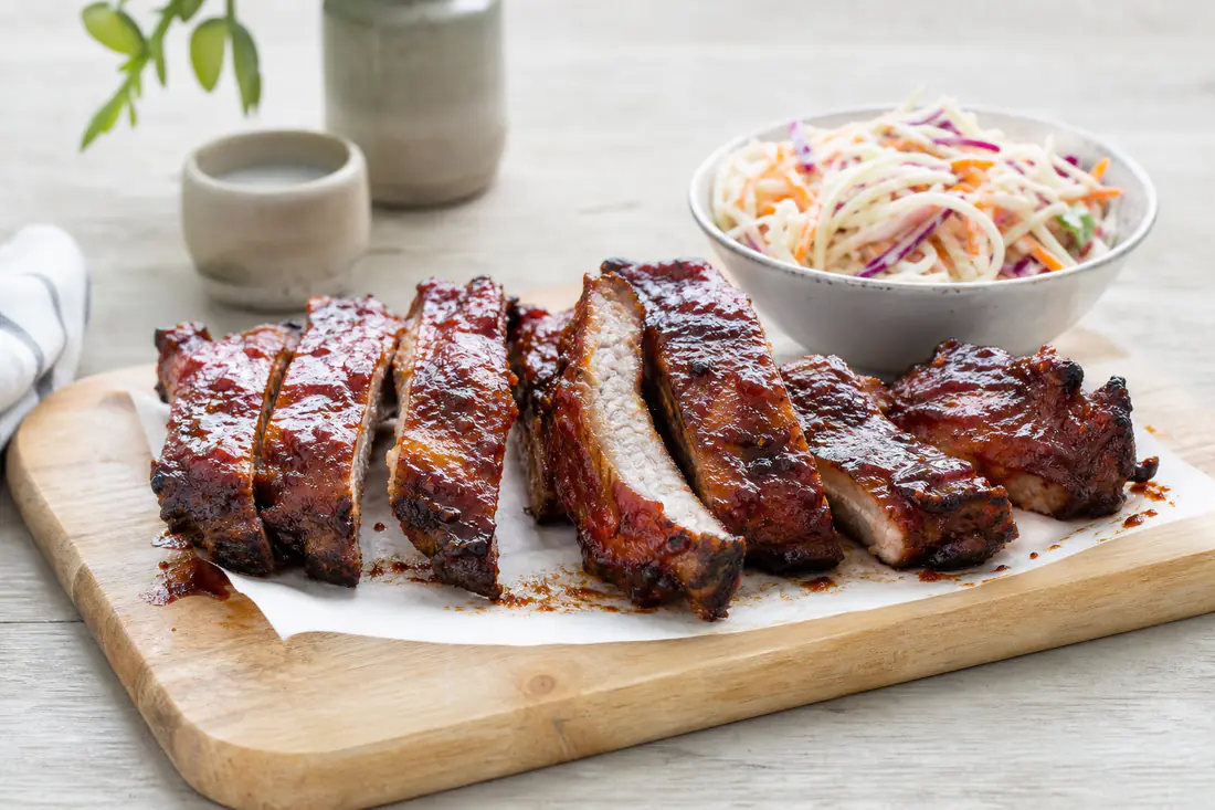 Glänzende Spareribs mit dunkler BBQ-Glasur, in einzelne Rippen geschnitten und auf einem Holzbrett angerichtet, dazu Coleslaw