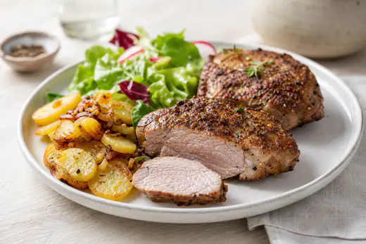 Zwei dicke Schweinenackensteaks mit gebräunter Gewürzkruste auf einem Teller, eines aufgeschnitten, mit Bratkartoffeln und grünem Salat