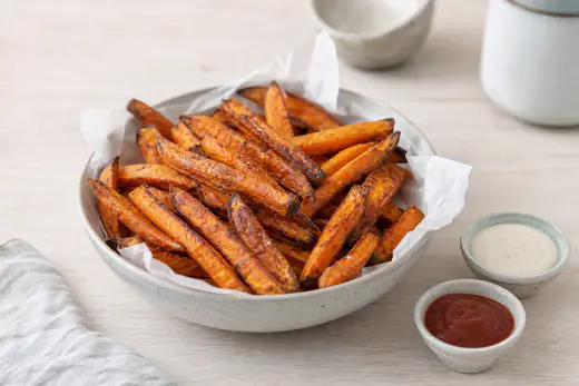 Knusprige orangefarbene Süßkartoffelpommes mit dunklen Rändern in einer mit Papier ausgelegten Schüssel, daneben zwei kleine Dips