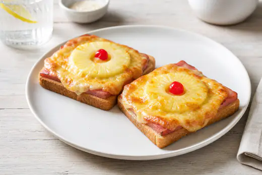 Zwei quadratische Toast Hawaii mit Schinken, Ananasring und goldbraun überbackenem Käse, in der Mitte je eine Cocktailkirsche, auf einem weißen Teller