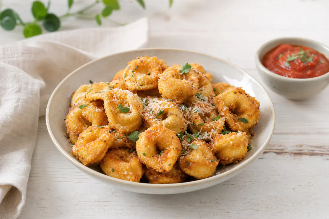 Knusprig gebackene goldbraune Tortellini mit Parmesan und Kräutern in einer Schale, daneben ein Schälchen Tomatendip