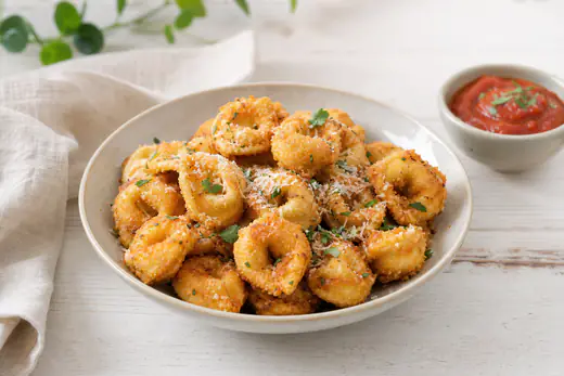 Knusprig gebackene goldbraune Tortellini mit Parmesan und Kräutern in einer Schale, daneben ein Schälchen Tomatendip