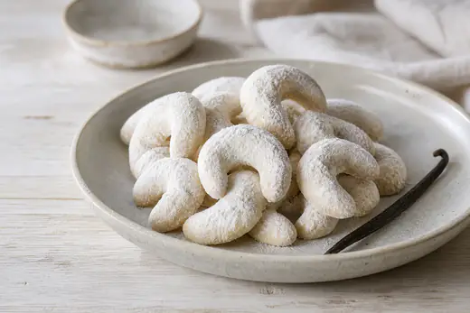 Hufeisenförmige Vanillekipferl dick mit Vanille-Puderzucker bestäubt auf einem Teller, daneben eine Vanilleschote
