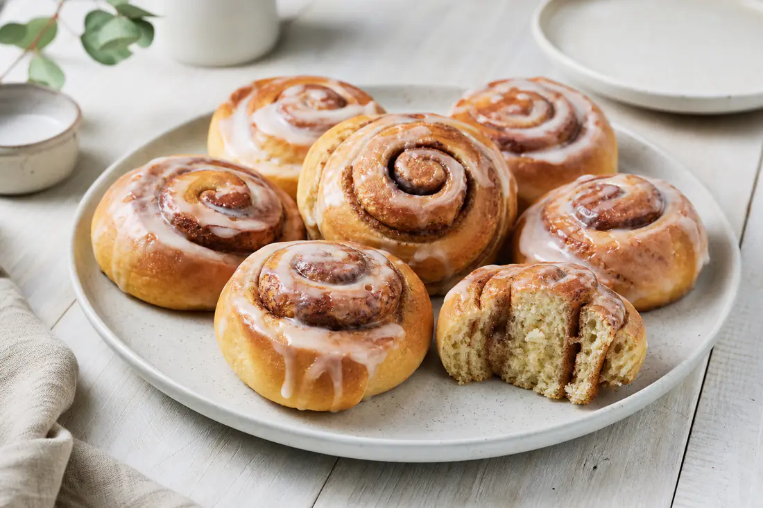 Goldbraune Zimtschnecken mit weißem Zuckerguss auf einem Teller, eine Schnecke angeschnitten mit Zimtspirale