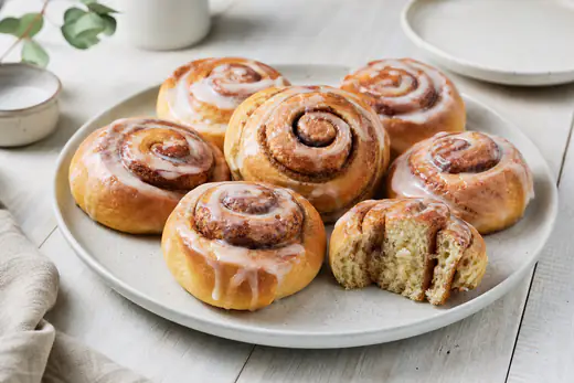 Goldbraune Zimtschnecken mit weißem Zuckerguss auf einem Teller, eine Schnecke angeschnitten mit Zimtspirale