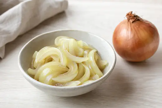 Weich gedünstete, glasige Zwiebelhalbringe in einer kleinen Keramikschale, daneben eine ganze gegarte Zwiebel mit Schale